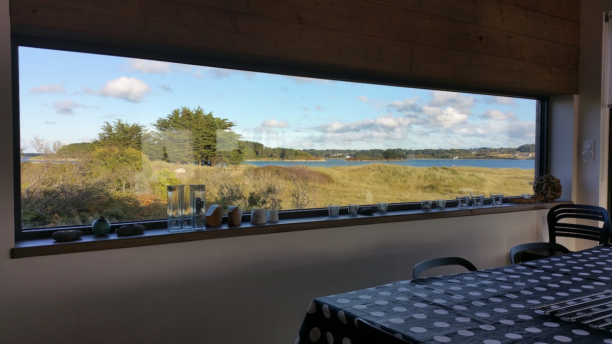 A large window offers a panoramic view of the surrounding landscape, featuring serene waters and lush greenery. A simple table with a polka dot tablecloth is visible in the foreground, and various decorative items are placed neatly along the window sill.