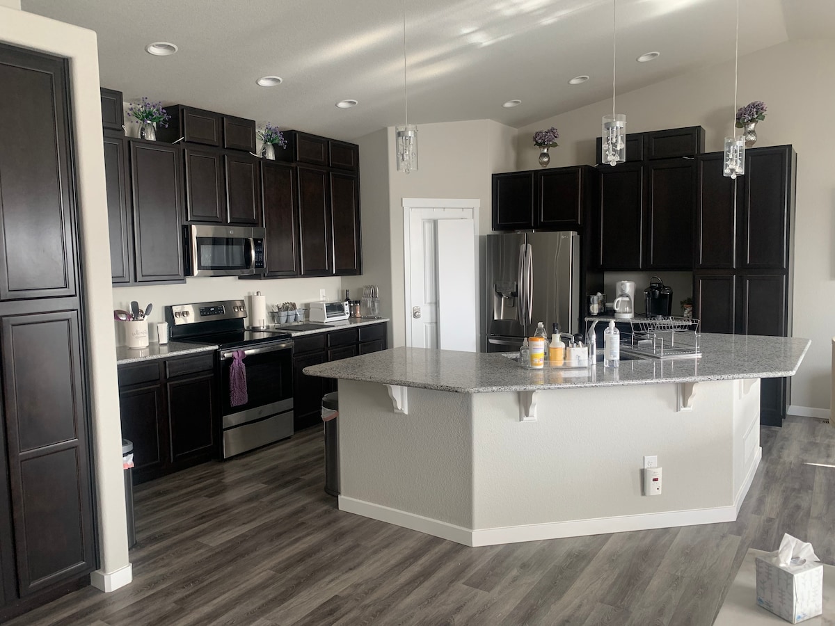 A modern kitchen features dark cabinetry and a large island with a granite countertop. Stainless steel appliances, including a refrigerator and microwave, are prominently displayed. Under-cabinet lighting enhances the work areas, while decorative elements add a touch of charm.