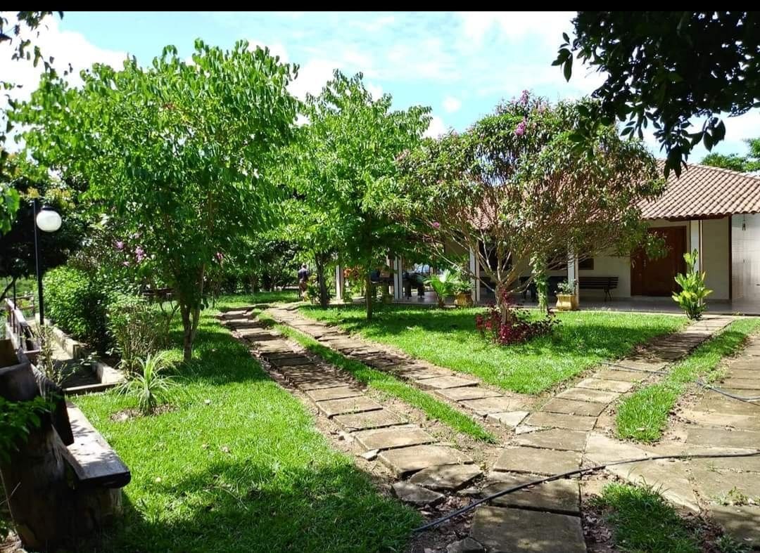 A spacious outdoor area is surrounded by lush greenery and colorful plants. Stone pathways lead through the garden, connecting various sections of the property, while trees provide shade. The charming house is visible in the background, featuring a tile roof and wooden doors.