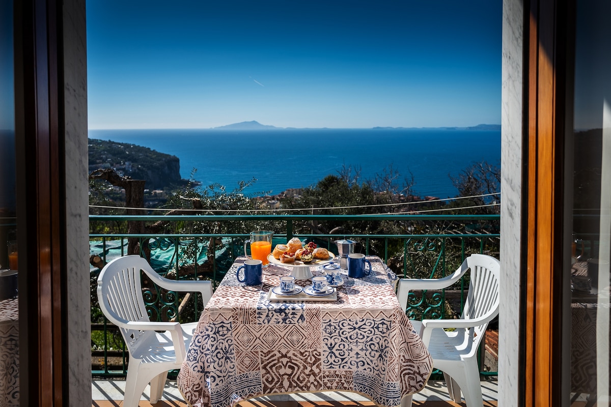 A dining table is set for breakfast on a balcony, featuring a view of the sea and distant islands. The table displays a spread of food, including pastries and beverages, complemented by white plastic chairs and vibrant blue accents.
