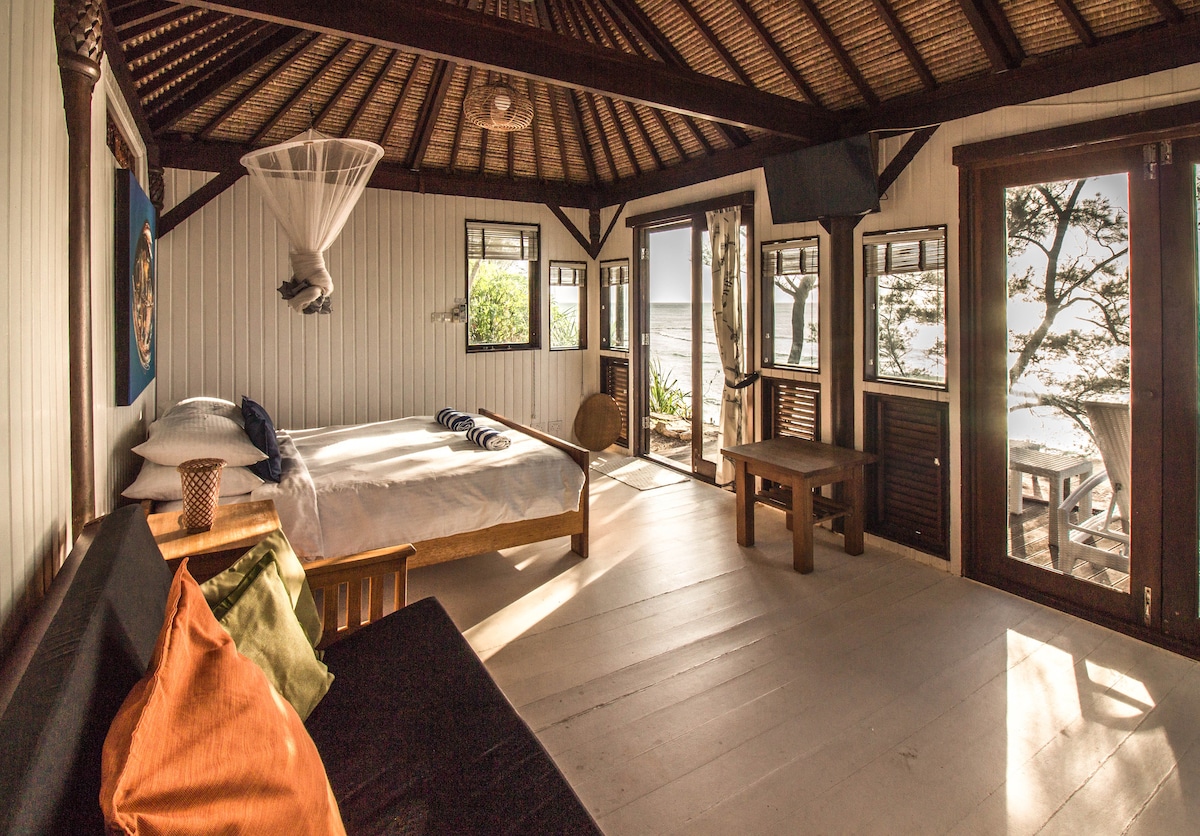 The spacious sleeping area features a large bed and a comfortable seating area with colorful cushions. Natural light enters through large windows, revealing views of the outdoor landscape. A soft mosquito net hangs above the bed, enhancing a sense of tranquility.