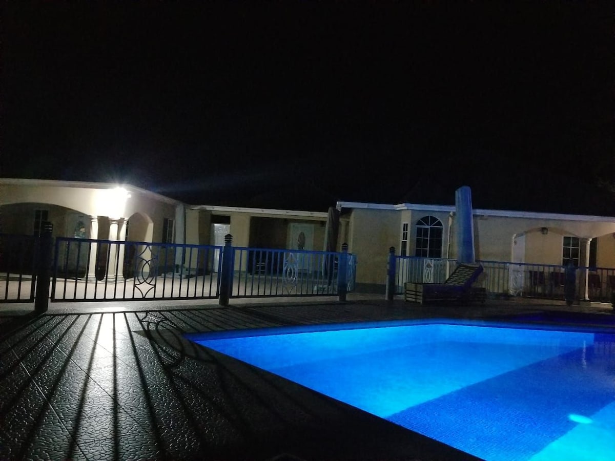 A spacious outdoor area is illuminated by soft lights, featuring a clear blue pool surrounded by a textured deck. A building is visible in the background, with multiple entry points and large windows. The serene atmosphere is heightened by the nighttime setting.