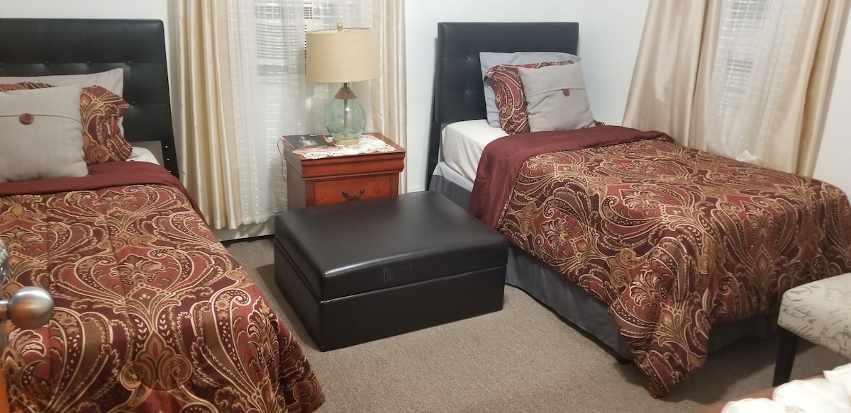 A cozy bedroom is featured with two beds dressed in patterned bedspreads. A bedside lamp provides soft lighting, while a black storage ottoman is positioned at the center. Light-colored drapes frame the windows, allowing natural light to enter the space.
