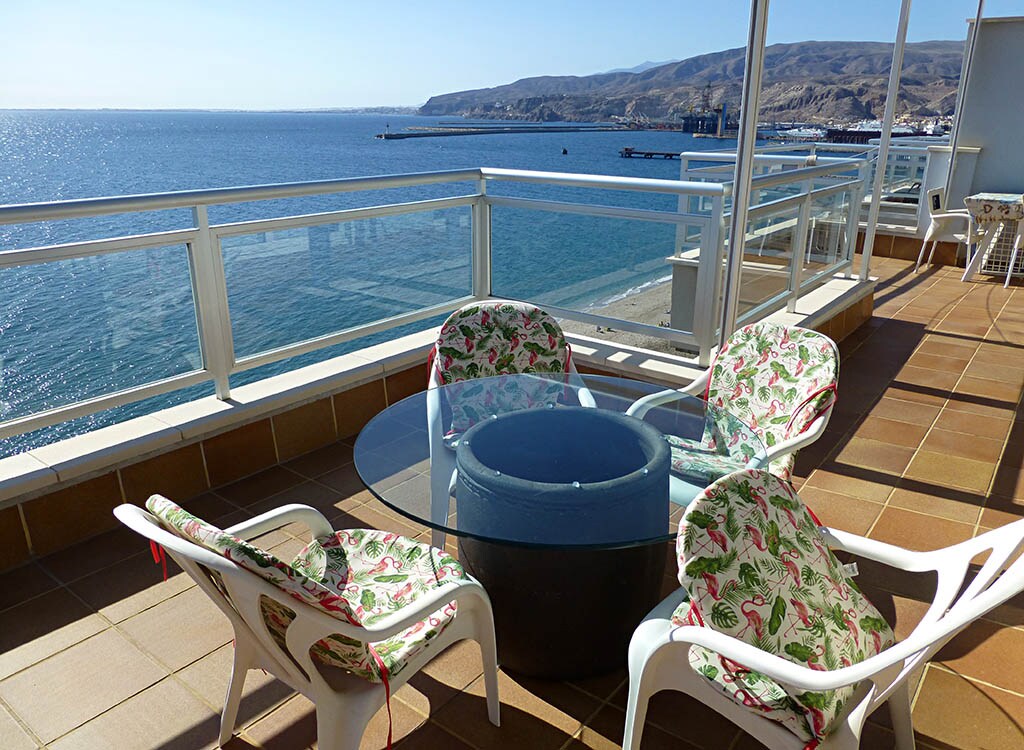 An outdoor terrace showcases a round glass table surrounded by four white chairs with colorful floral cushions. The expansive view features the ocean and distant hills under a clear sky.