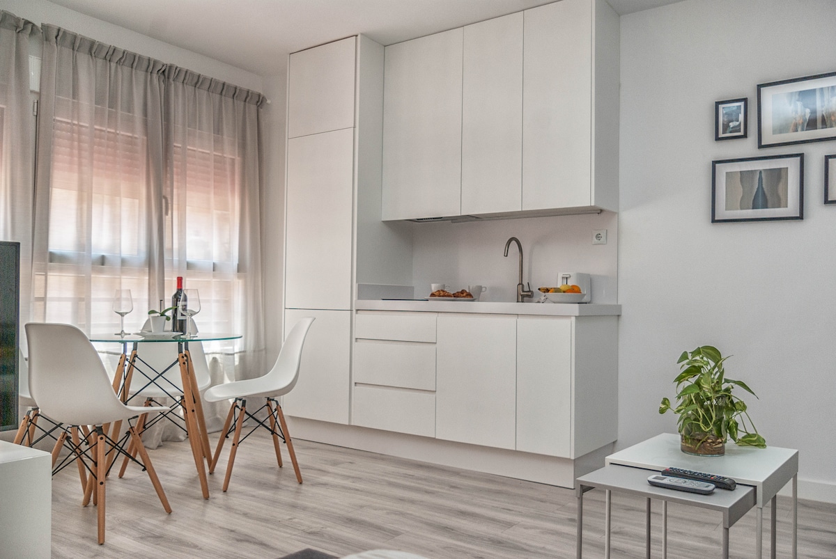 The kitchen area features minimalist white cabinetry and a sleek countertop. A glass dining table is surrounded by four modern chairs with wooden legs. Natural light filters through sheer curtains, enhancing the light wood flooring and a small potted plant on the coffee table.