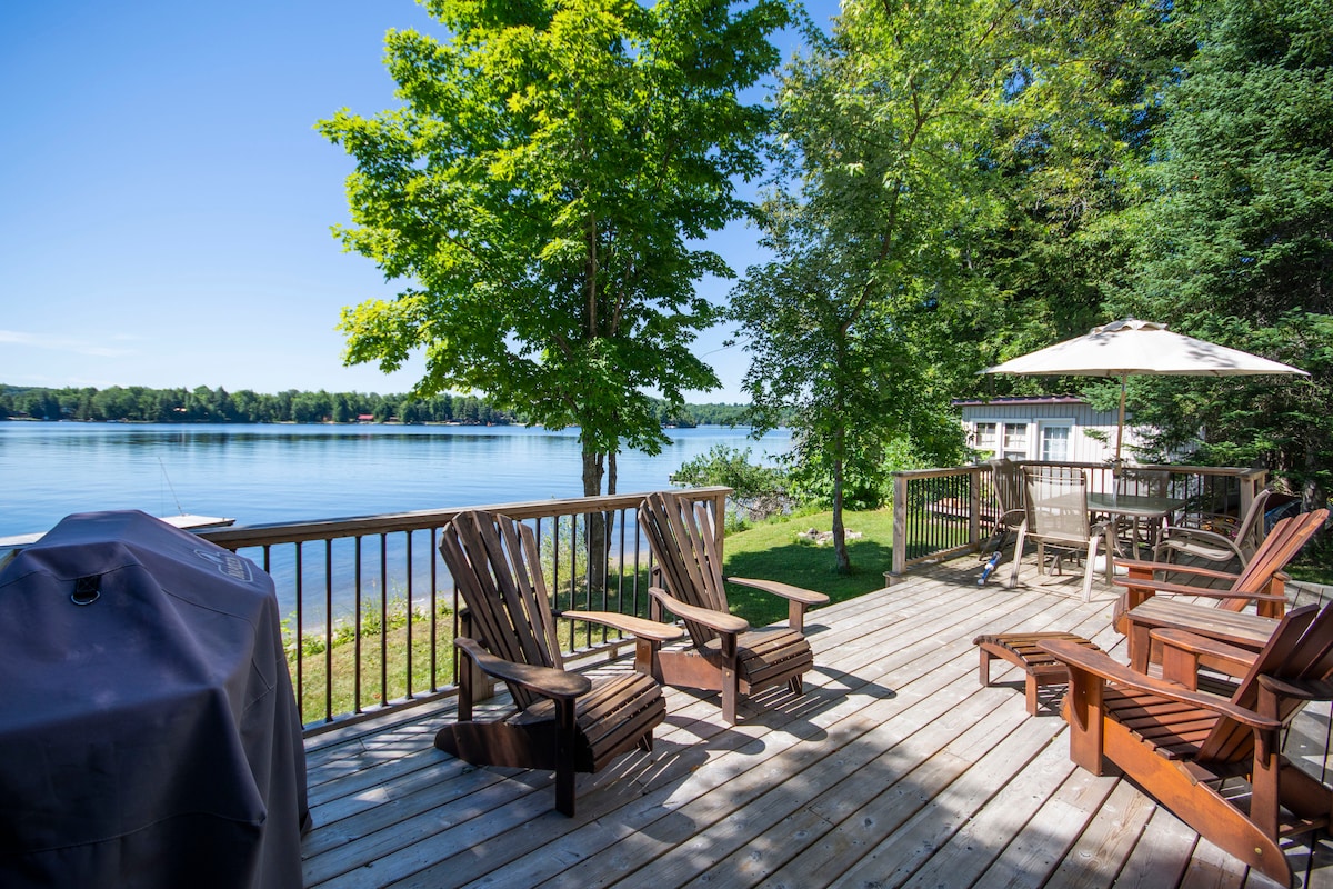 A spacious deck overlooks Horseshoe Lake, featuring wooden Adirondack chairs and a patio table with an umbrella. A propane BBQ is positioned near the railing, surrounded by lush greenery. The calm water reflects the blue sky, creating a peaceful outdoor setting.