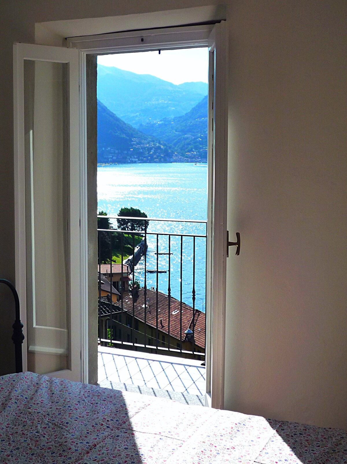 A bright doorway opens to a balcony, offering a view of Lake Como and the surrounding mountains. Sunlight reflects on the water, creating a serene atmosphere. The floral-patterned fabric of the bed frame is partially visible, adding a touch of comfort to the space.
