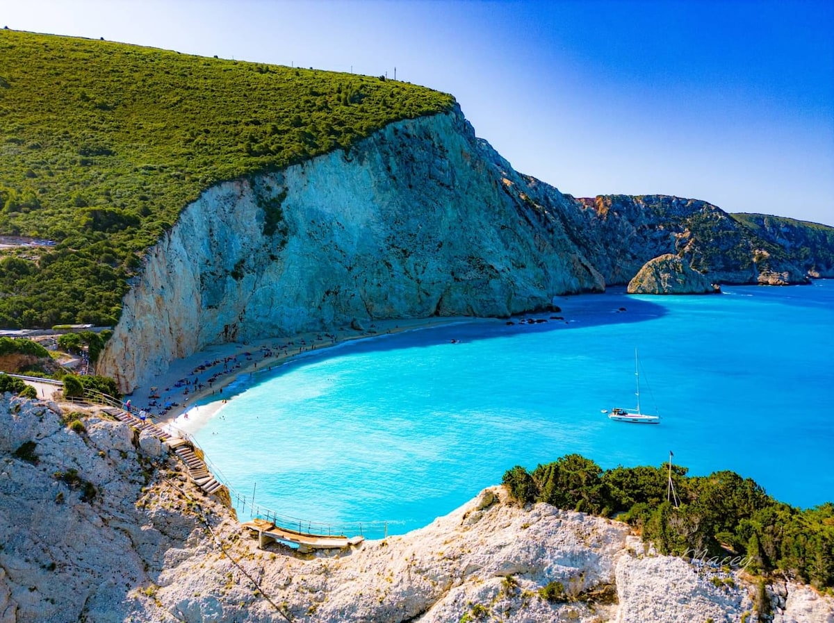 A scenic coastline is displayed with a sandy beach nestled between rocky cliffs. The vibrant blue waters are viewed, along with a small sailboat floating in the calm sea. Lush green hills rise in the background, creating a serene natural environment.