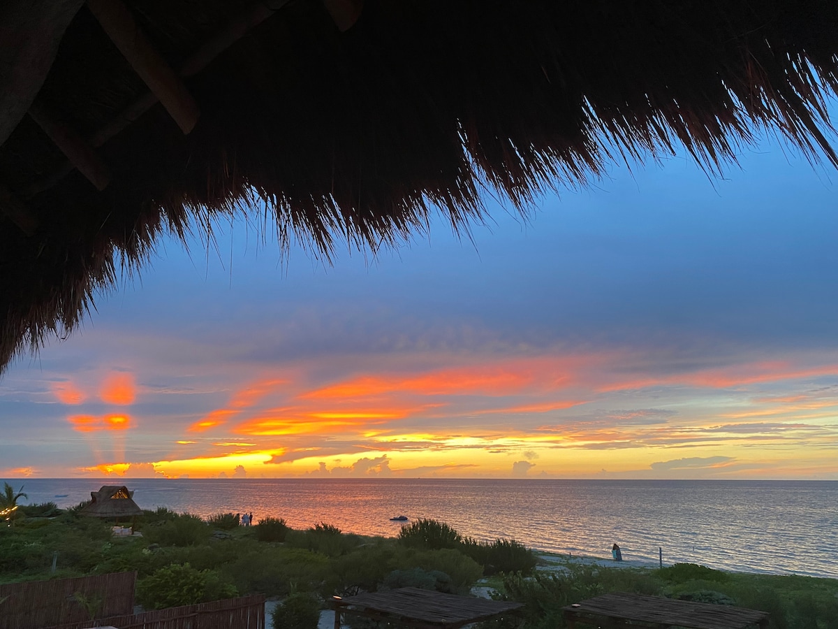 A vibrant sunset casts an array of colors across the sky, blending shades of orange and blue. The expansive view of the ocean is framed by tropical greenery, with distant silhouettes of a thatched-roof structure and beachgoers visible along the shoreline.