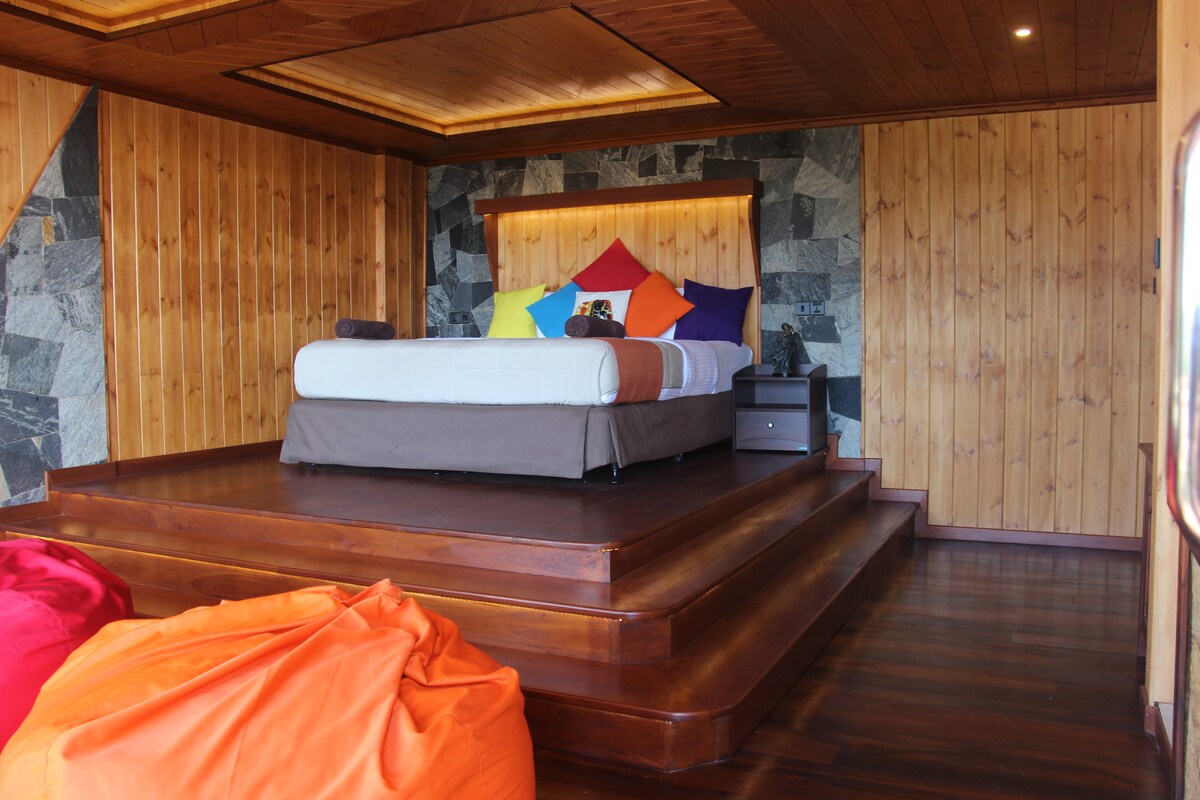 A cozy room features a spacious bed adorned with colorful pillows resting against a stone accent wall. Soft wood paneling covers the walls and floor, while two large beanbags in vibrant hues provide additional seating options. Warm lighting creates a welcoming ambiance.
