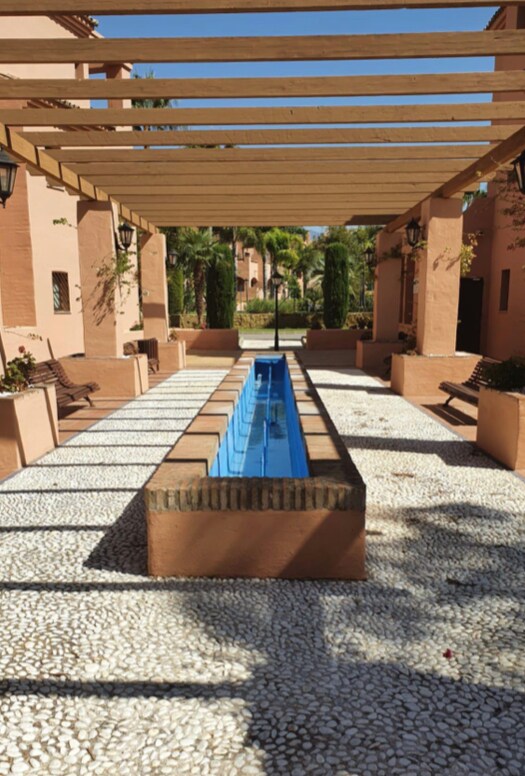 A tranquil outdoor area is presented with a long, narrow water feature running through the center. Wooden benches are placed along the sides, and the ground is covered with white pebbles, creating a serene atmosphere enhanced by surrounding greenery and shaded pergolas.