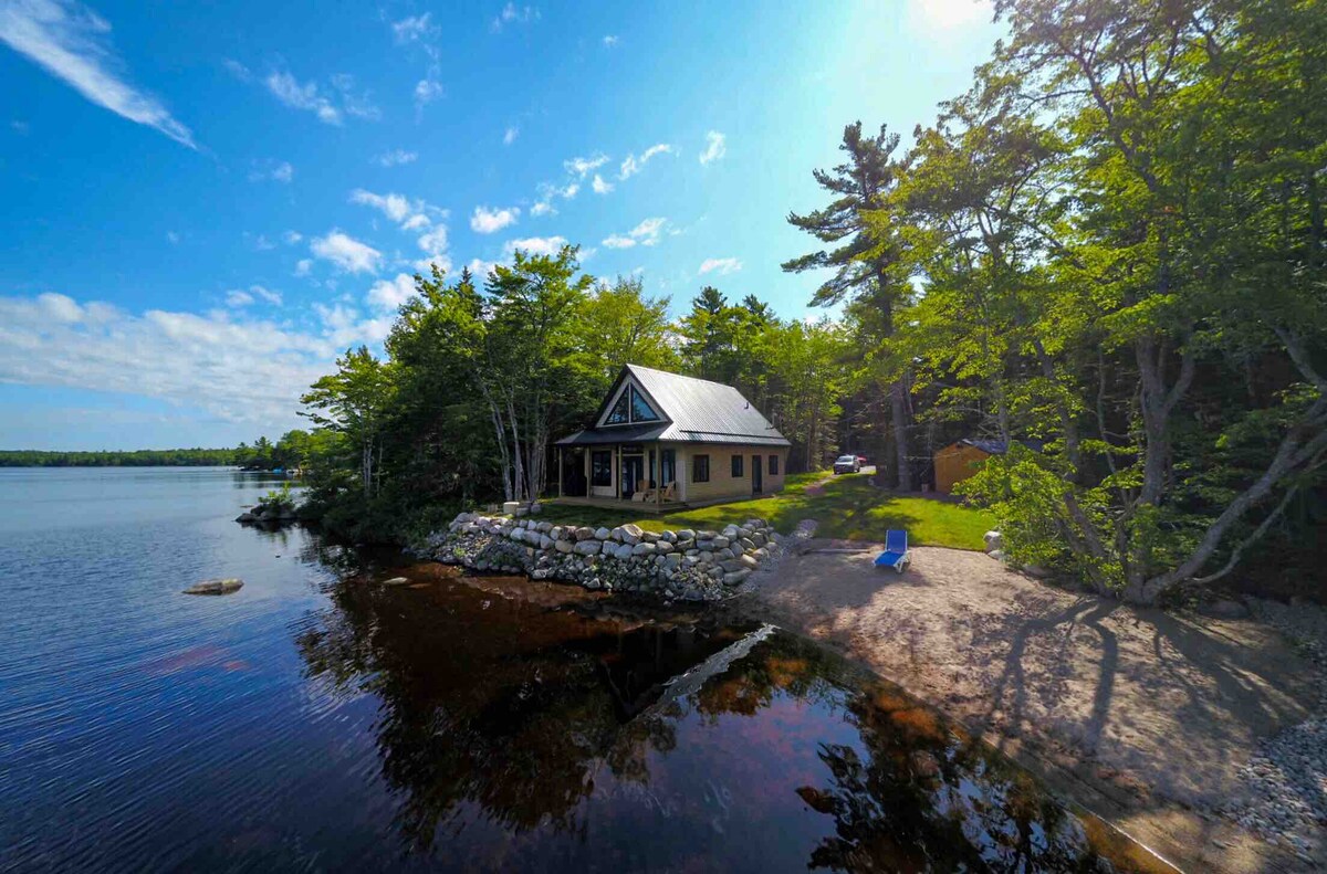 The cottage is viewed from above, bordered by trees along the shoreline of Grand Lake. A sandy beach is visible, flowing into calm water reflecting the clear blue sky. The structure features a sloped roof and a welcoming front porch, blending harmoniously with the natural landscape.