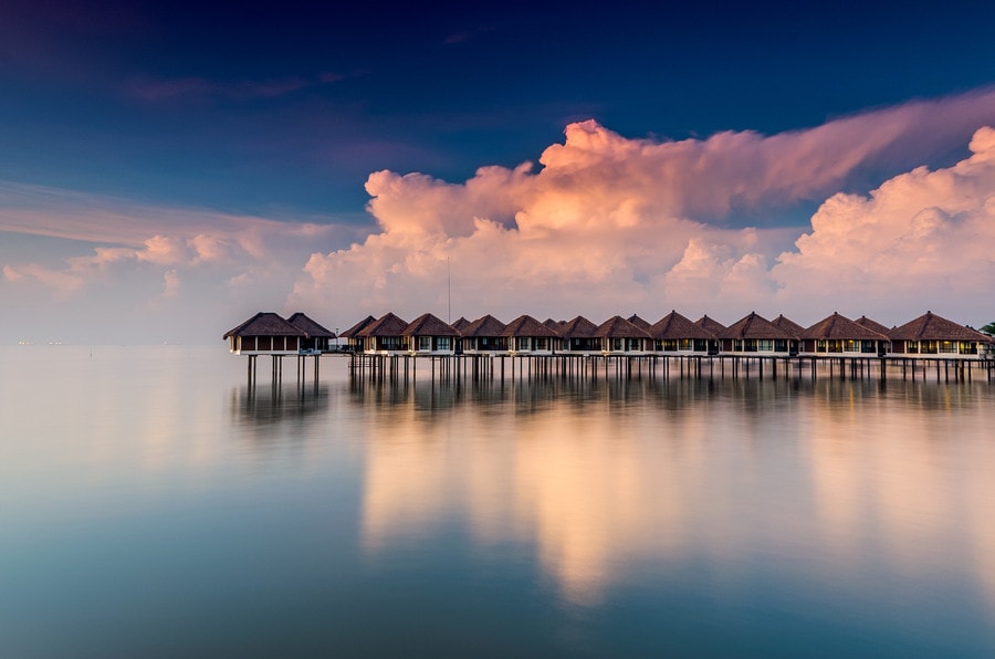 A series of overwater bungalows is positioned atop stilts, extending into calm waters. The structure features a thatched roof design, reflecting softly on the water's surface. A vibrant sky filled with clouds is observed in the background, creating a serene coastal atmosphere.