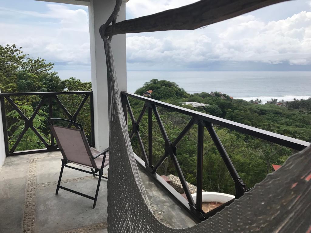 A balcony area features a hammock and a minimalist chair, offering a relaxing space to enjoy the surroundings. Lush greenery frames the view of the ocean and distant hills under a cloudy sky, enhancing the tranquil atmosphere.