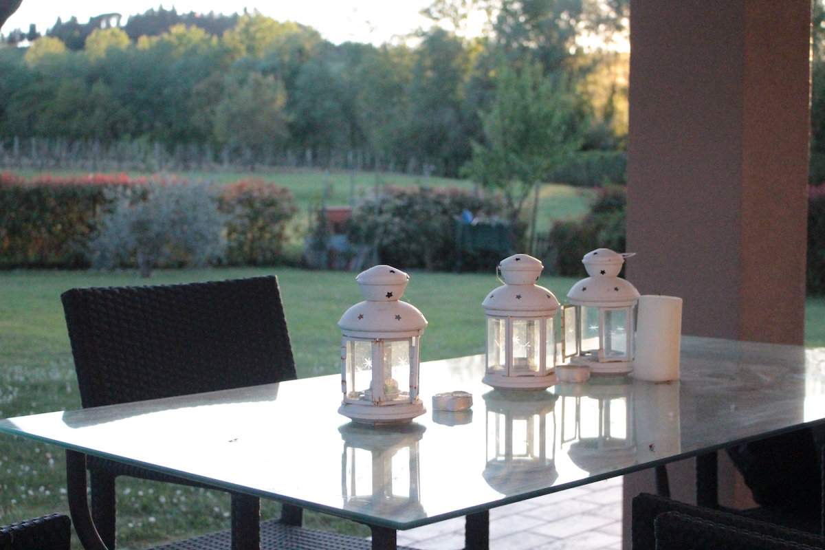 A dining table is set outdoors, featuring three white lanterns and a candle on its glass surface. Surrounding the table are dark wicker chairs, while a lush landscape with trees and hedges can be seen in the background, offering a serene countryside view.