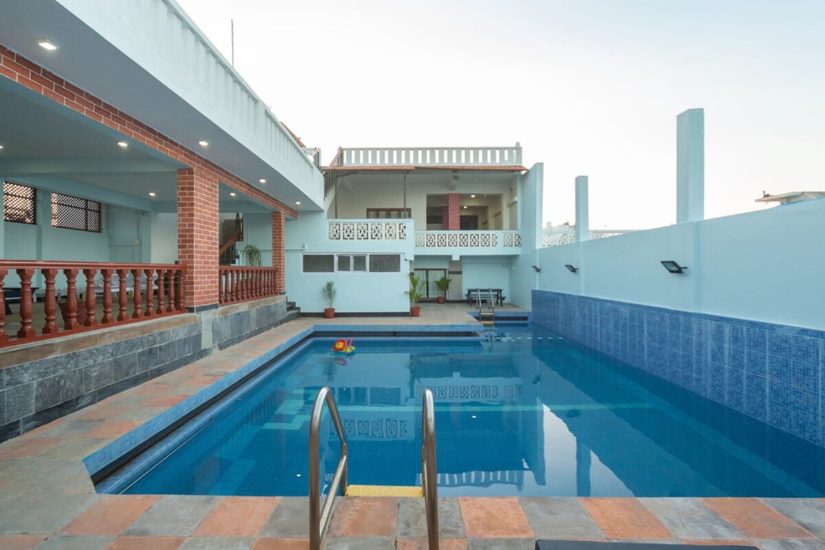 A swimming pool is featured with a clear blue surface reflecting the sky. Surrounding the pool, a spacious area includes lounge chairs and a brick terrace. The building's exterior is visible in the background, showcasing a balcony and windows that overlook the pool area.