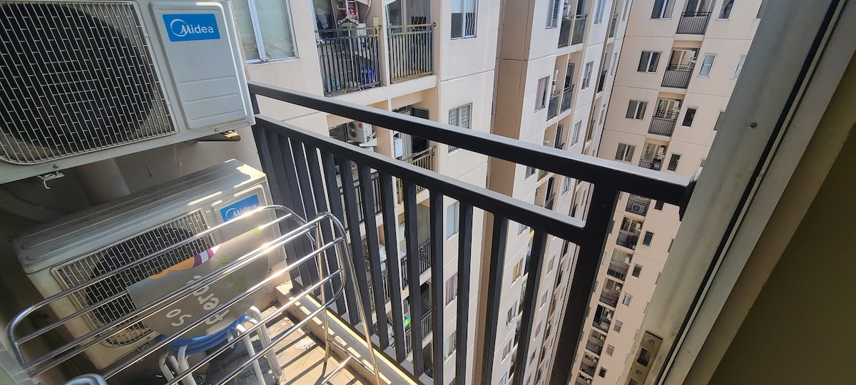 A balcony view showcases a metal railing overlooking a courtyard enclosed by tall residential buildings. An air conditioning unit is mounted to the left, and a drying rack is positioned on the balcony floor.