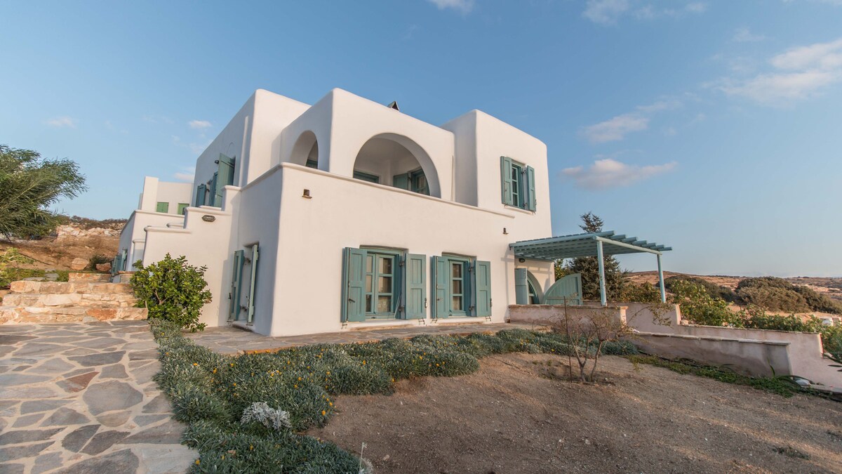 The exterior of the villa is showcased with whitewashed walls and teal wooden shutters. An arched entrance is visible, along with a covered terrace featuring a pergola. A stone pathway leads through a landscaped area with local vegetation and flowering plants, all under a clear sky.