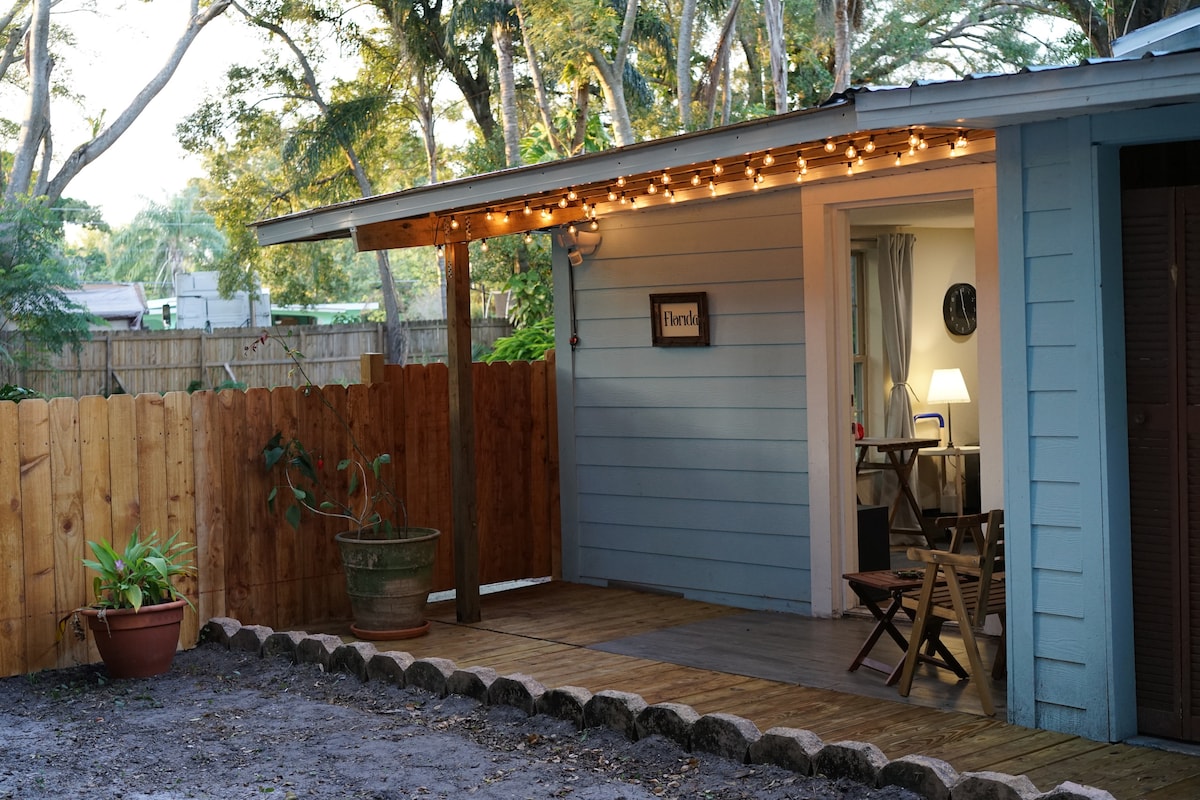 A small, covered porch features string lights hanging overhead, offering a cozy and inviting space. The entrance is framed by a wooden fence, and simple outdoor furniture is placed on the wooden decking, enhancing the serene outdoor setting among the trees.