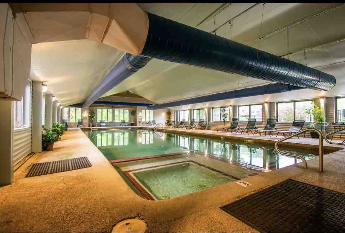 An indoor pool area features a large swimming pool with a separate hot tub. Surrounding the pool, comfortable lounge chairs are positioned along the spacious deck. Large windows provide natural light, creating a bright and inviting environment.