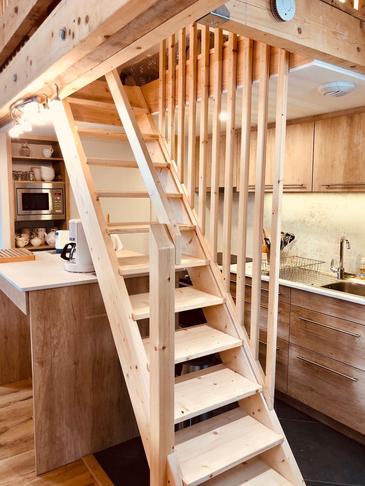 A wooden staircase with vertical railings leads to the mezzanine level, connecting the living area and kitchen. The kitchen is equipped with modern appliances and cabinets, complemented by warm wood tones and natural light from the nearby window.