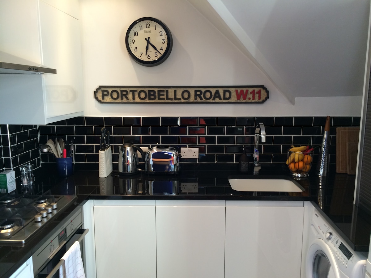 The kitchen features modern, high-gloss white cabinetry contrasted by black tile backsplash. A black countertop holds a kettle, toaster, and a bowl of fresh fruit. A vintage-style clock is mounted on the wall above, while stainless steel appliances and a washing machine are visible.