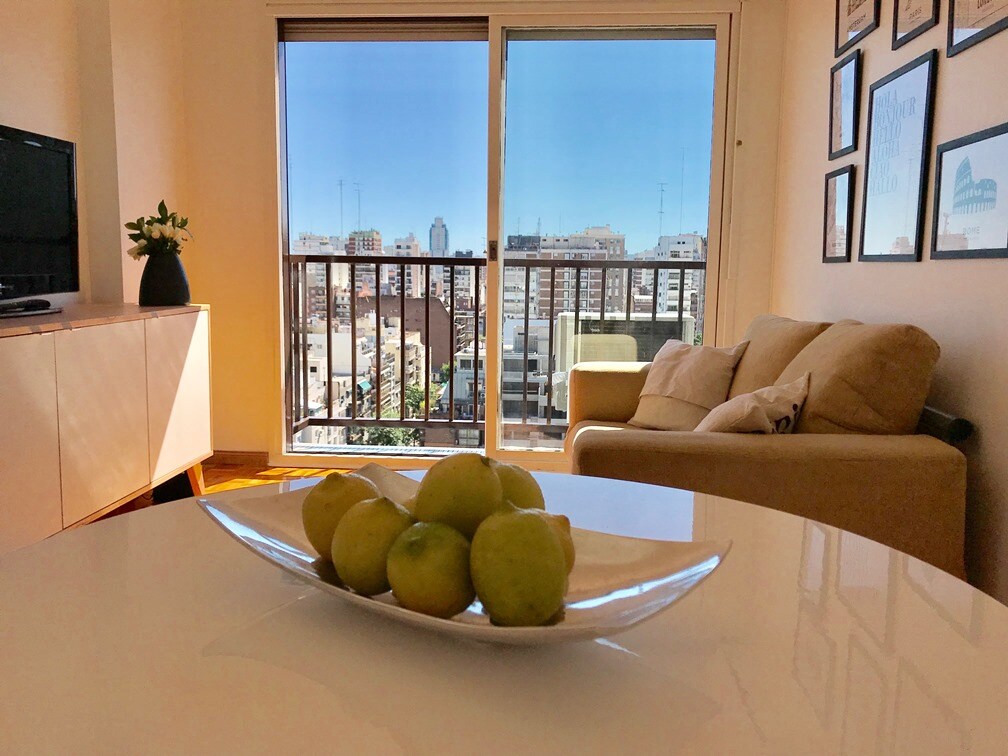 A living space is displayed, featuring a light-filled atmosphere. A white dining table holds a bowl of green apples. A comfortable sofa is positioned near large windows, offering an unobstructed view of the city skyline. Framed artwork adorns the adjacent wall.