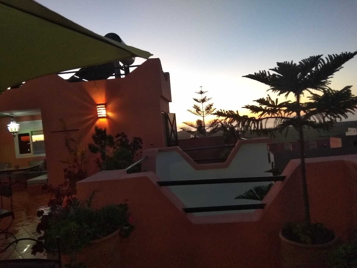 The rooftop terrace showcases a view of the horizon during dusk, with soft colors blending in the sky. Potted plants line the edges, while a shaded seating area invites relaxation. Ambient lighting adds a gentle glow to the surrounding walls, enhancing the serene atmosphere.
