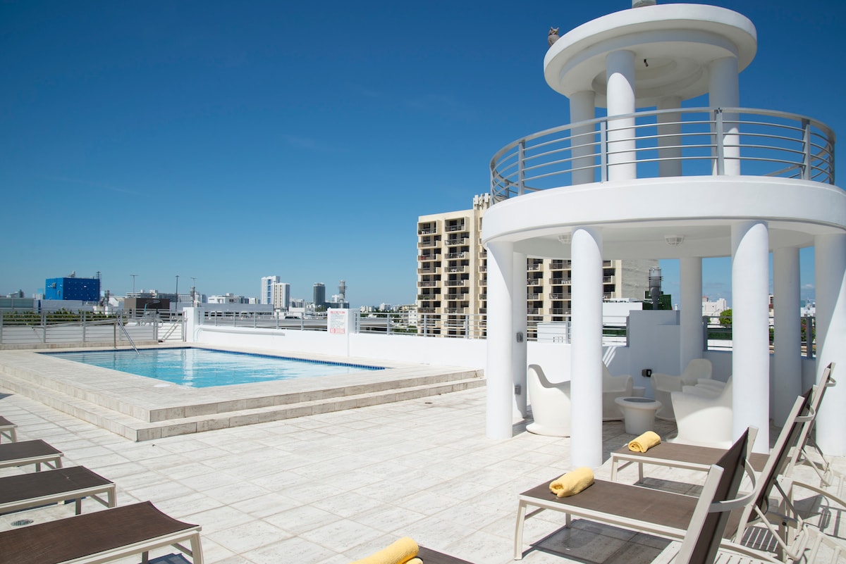 A spacious rooftop pool area features a clean, blue swimming pool surrounded by lounge chairs. A white decorative structure with an open circular design is present, providing shade. The backdrop includes city buildings and a clear blue sky, enhancing the open atmosphere.