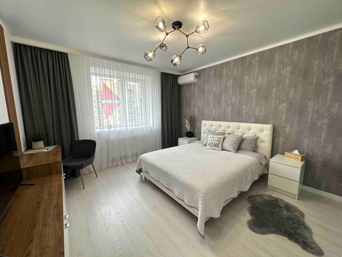 A well-lit bedroom features a spacious bed with a textured duvet positioned against a grey accent wall. A comfortable chair is placed near the window, which is dressed in dark curtains, allowing natural light to filter through. A small bedside cabinet is situated beside the bed.
