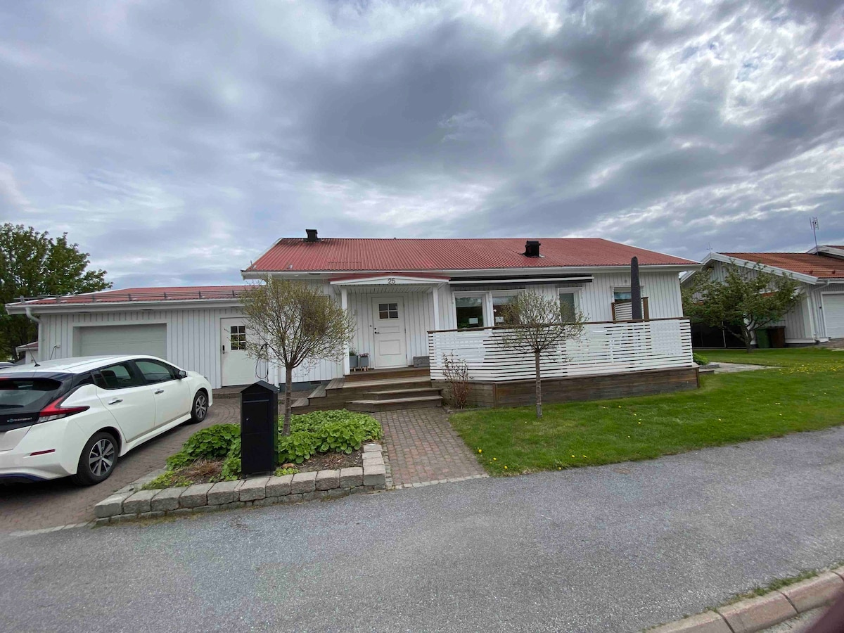 A single-story villa is depicted with a red roof and white wooden exterior. A paved walkway leads to the entrance, bordered by well-maintained grass and small trees. A vehicle is parked in the driveway, indicating convenient access.
