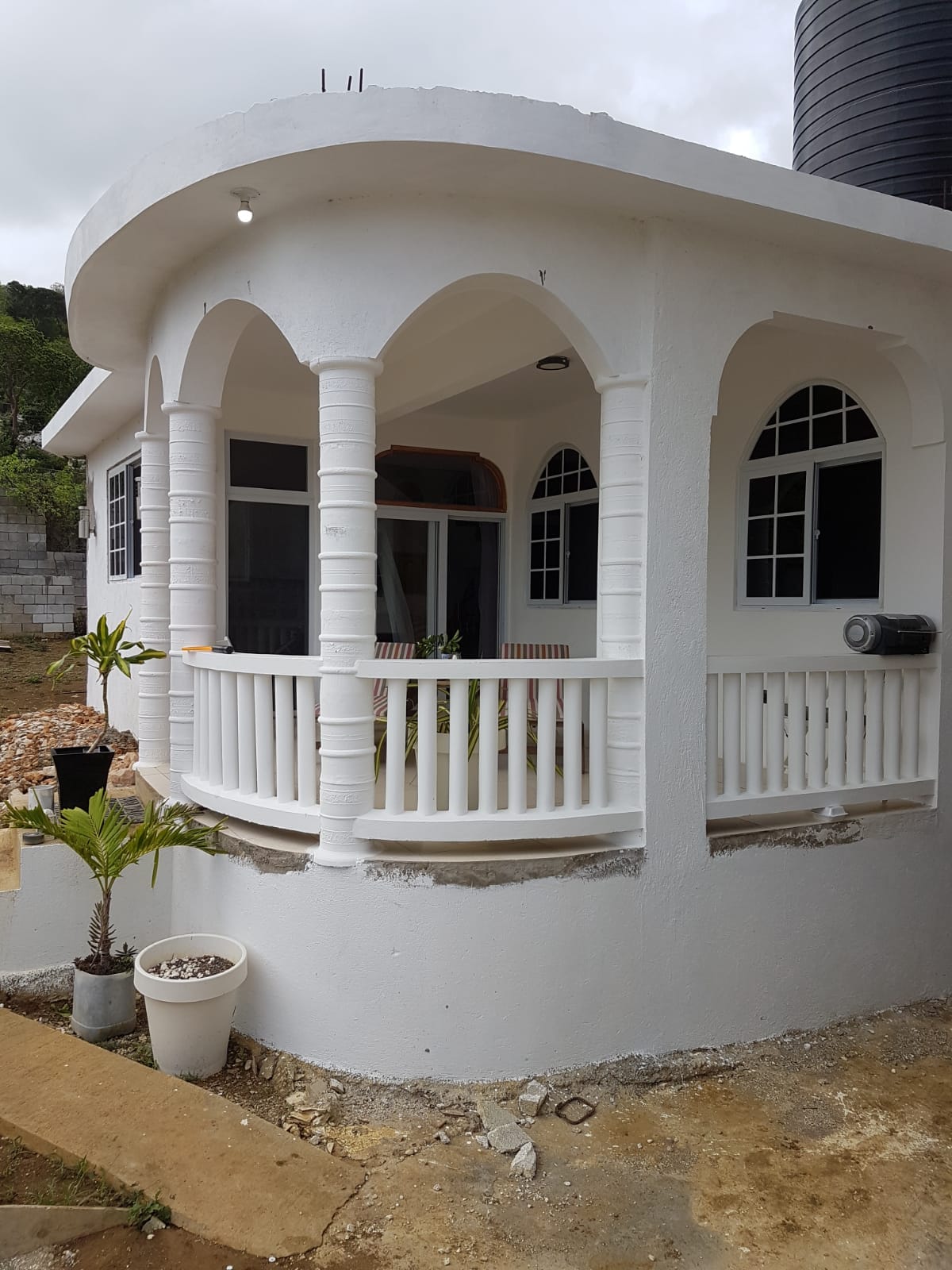 A corner of the villa is shown, featuring a white exterior and curved balcony adorned with white railings. Large windows, framed in black, allow ample light inside. Lush greenery is positioned in a planter near the entrance, enhancing the serene atmosphere of the property.