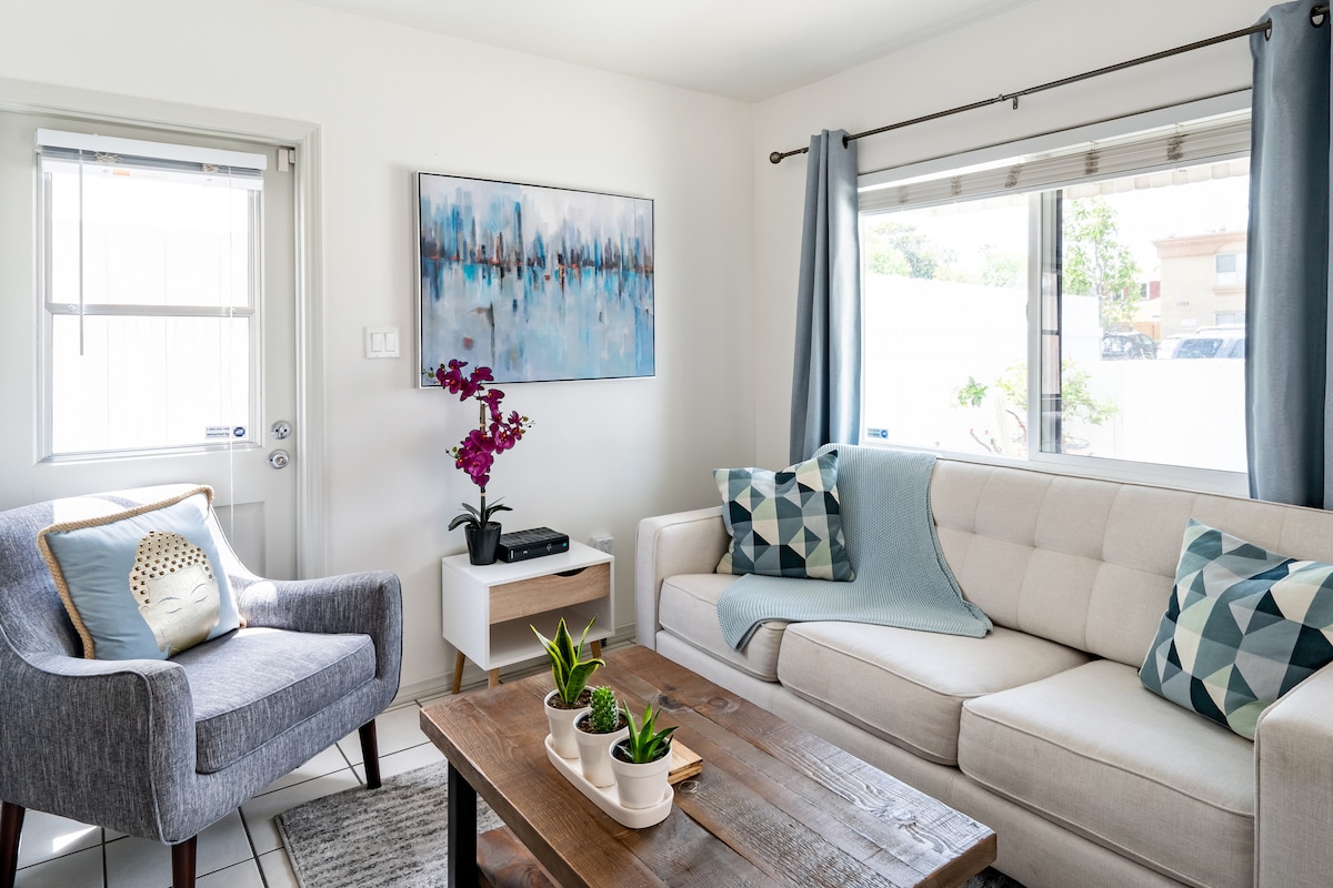 A light-filled living room features a cozy sectional sofa adorned with decorative cushions and a soft throw. A comfortable armchair sits beside a small side table with an orchid. Natural light streams through the large window, enhancing the inviting atmosphere.