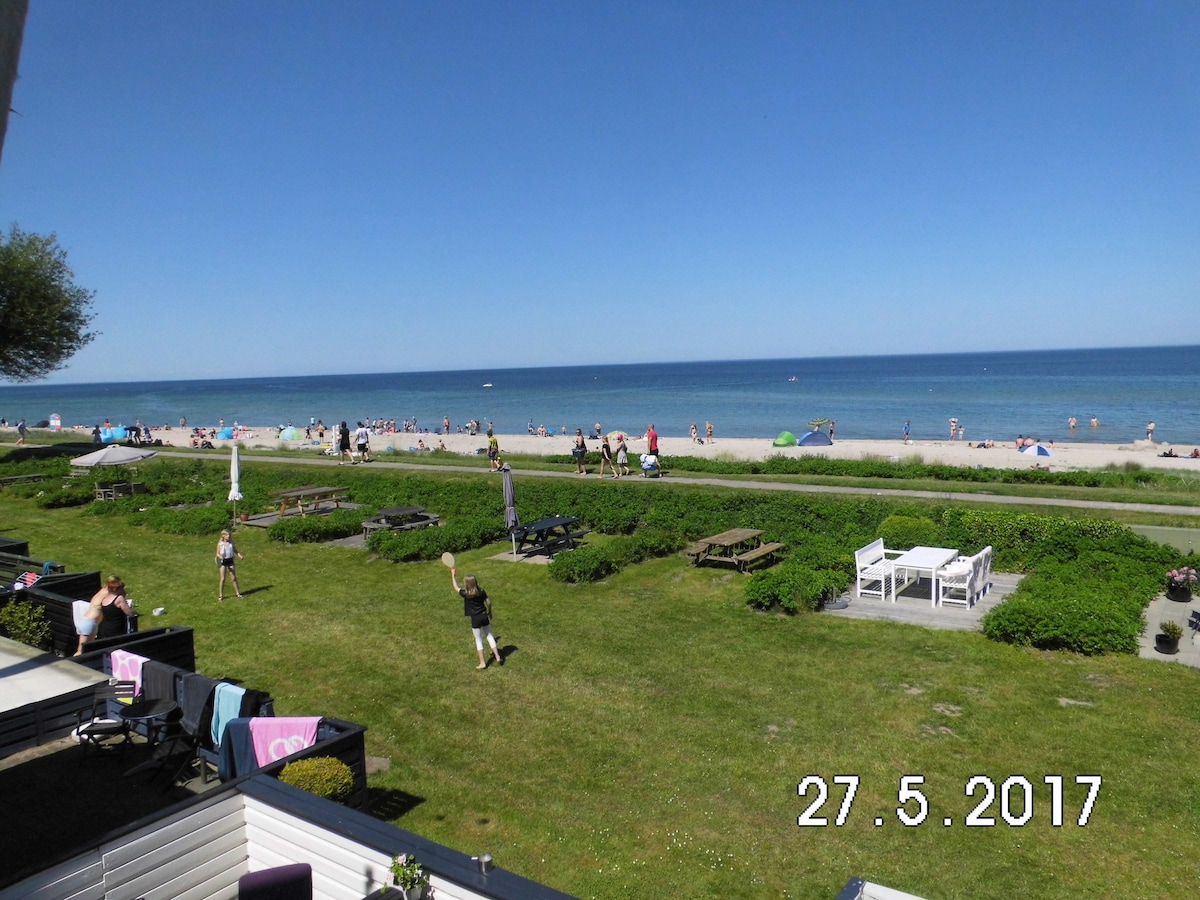 A view of a sandy beach lined with sunbathers and colorful umbrellas extends along the coastline. In the foreground, a well-maintained grassy area features picnic tables and a pathway leading to the beach. The clear blue sky complements the tranquil sea.