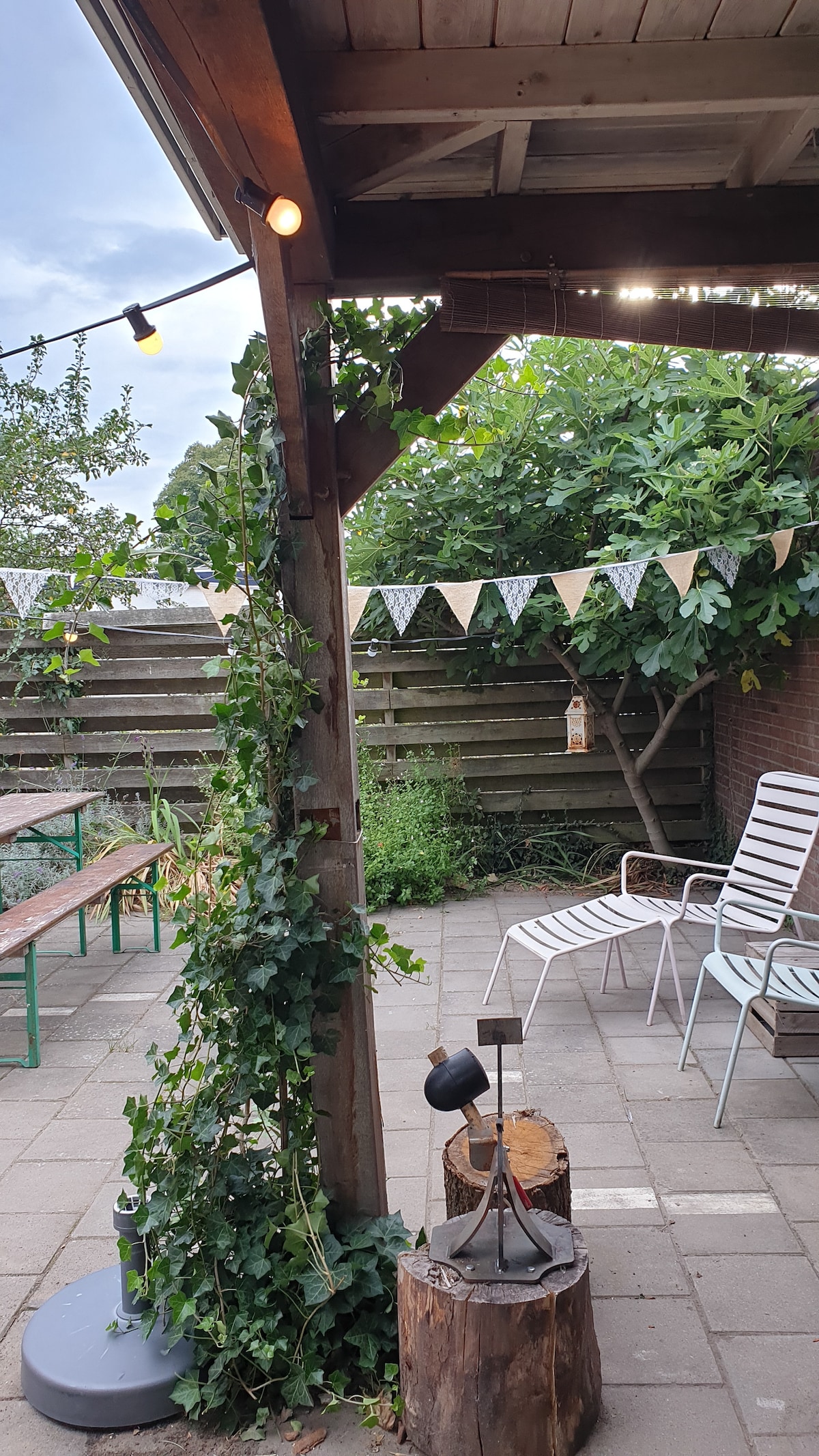 An outdoor patio space is presented with a decorative garland strung between wooden beams. Greenery is climbing one post, with a rustic wooden stump nearby. Metal lounge chairs are arranged for seating under ambient lighting, surrounded by a neatly manicured garden and wooden fencing.