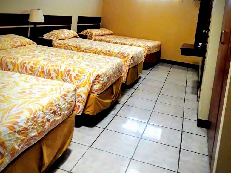 The room features four double beds, each covered with brightly patterned bedding in warm hues. A small table with a lamp is visible, along with a tiled floor that enhances the spaciousness. The walls are painted a light color, contributing to a well-lit environment.