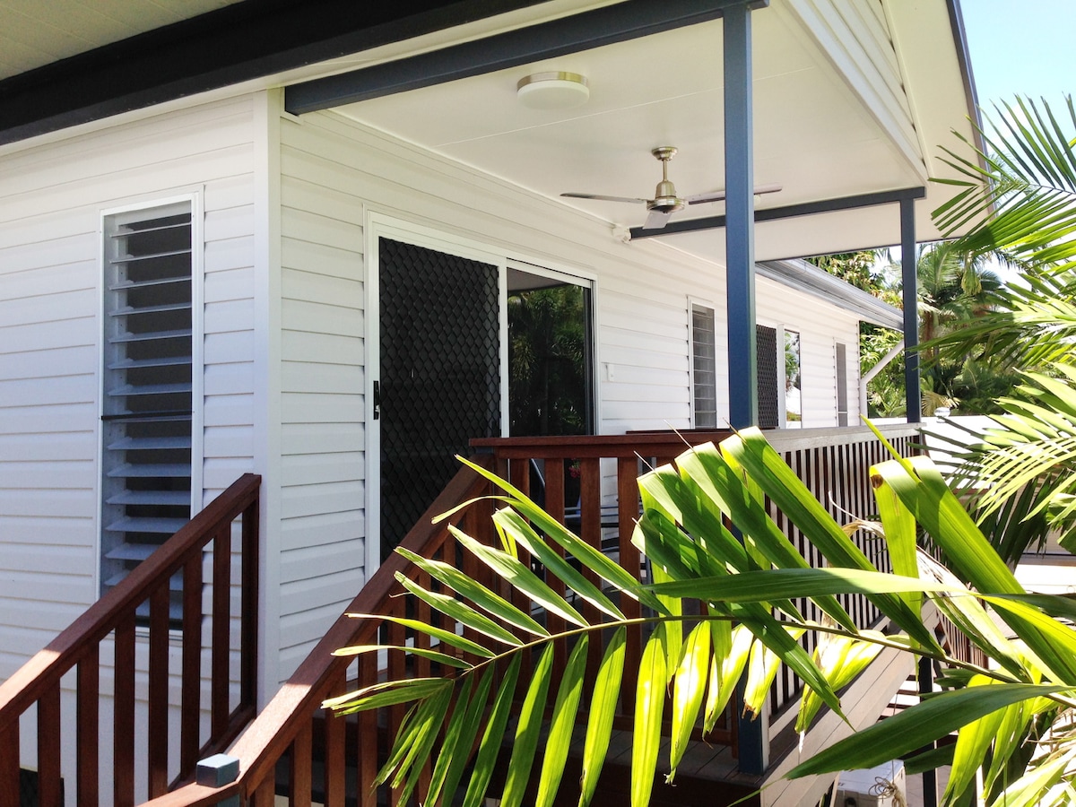 The exterior of the Granny Flat is visible, featuring a bright white facade accented by large windows and a wooden deck. Lush green palm fronds frame the image, suggesting a serene environment. A ceiling fan can be seen mounted above the porch area.