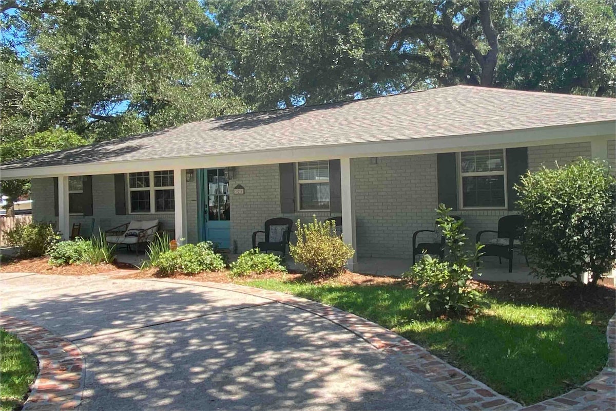 The exterior of the home features a welcoming façade with light-colored brick and a shingled roof. A landscaped front yard contains greenery and gravel paths, while inviting seating areas are positioned on the porch. Sunlight filters through the trees, creating dappled shadows on the ground.