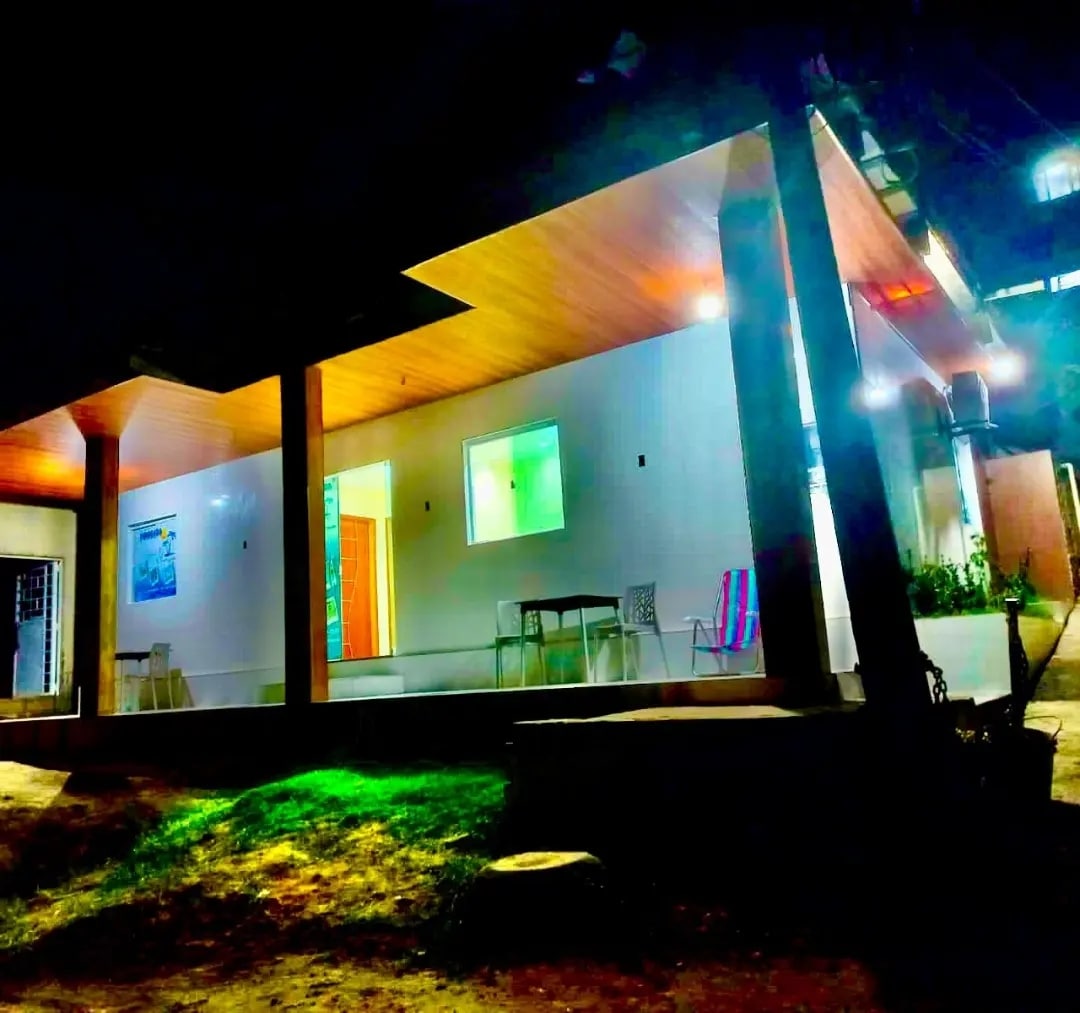 The exterior of a charming house is illuminated at night, revealing a spacious porch with seating arrangements. Brightly lit windows highlight the inviting interior, while soft grass patches enhance the surrounding area, providing a sense of tranquility.
