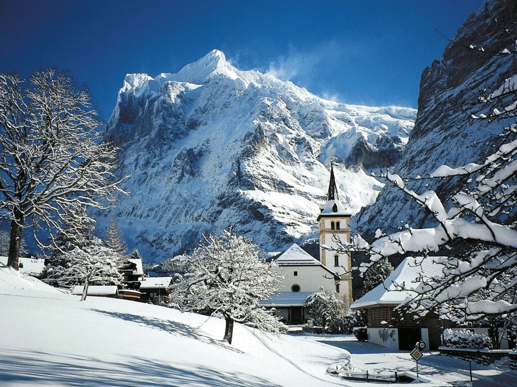 A serene winter landscape features a quaint village with a church, surrounded by snow-covered trees. Majestic mountains rise in the background, showcasing a pristine blue sky and a blanket of snow covering the ground.