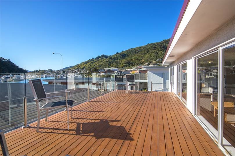 A spacious wooden deck is visible, equipped with outdoor seating for relaxation. The area overlooks a marina filled with boats and is framed by lush green hills in the background, all under a clear blue sky.