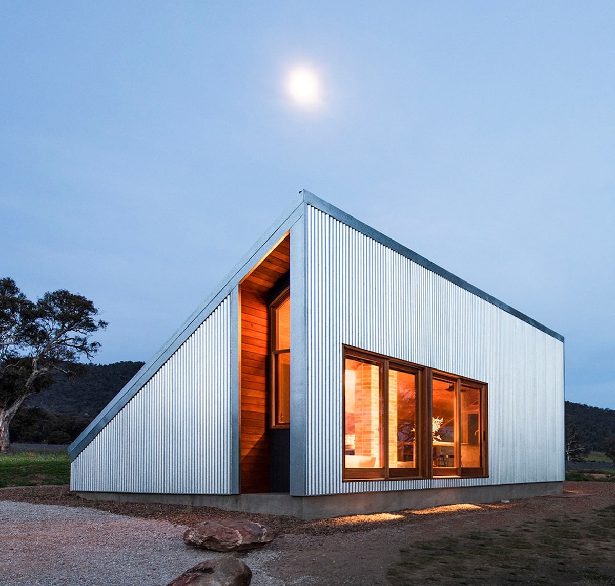 A Cameron Anderson Architects home sits below a full moon. Its windows and large open doorway show a warm, wooden interior as the corrugated metal ridges of its outer walls reflect the moonlight.