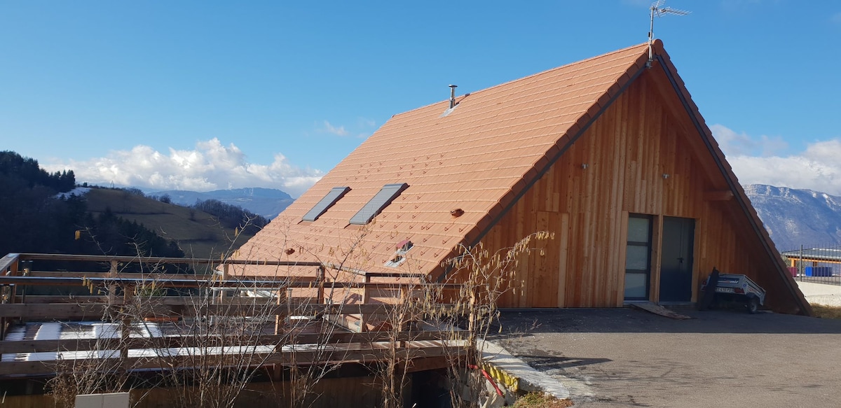 An A-frame building is showcased with a sloped wooden roof and large windows on either side. The entrance features a deck area with a railing, positioned against a backdrop of rolling hills and a clear blue sky, highlighting the natural surroundings.