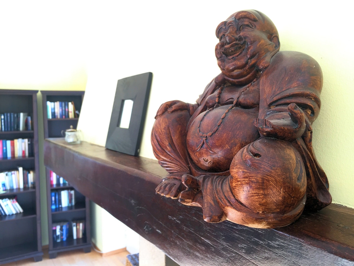 A wooden Buddha statue is positioned on a dark wooden mantel. The figure has a joyful expression and is surrounded by soft lighting. In the background, a bookshelf filled with various books is visible, contributing to a serene atmosphere.