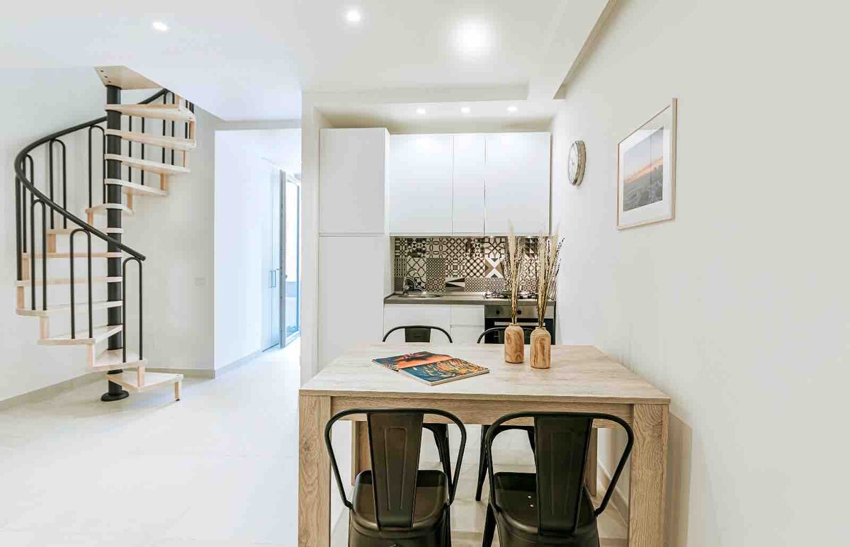 A modern kitchen area is featured, with a dining table surrounded by black chairs. A spiral staircase leads to the upper levels. The kitchen has white cabinetry and patterned tile backsplash, complementing the overall bright and airy space.