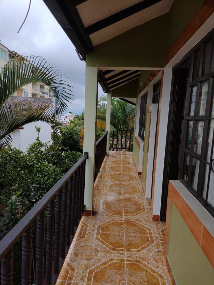A covered balcony is showcased, featuring a tiled floor with intricate patterns. Lush green plants and palm trees are visible in the background, enhancing the tranquil ambiance. The structure's warm exterior colors complement the inviting outdoor space.
