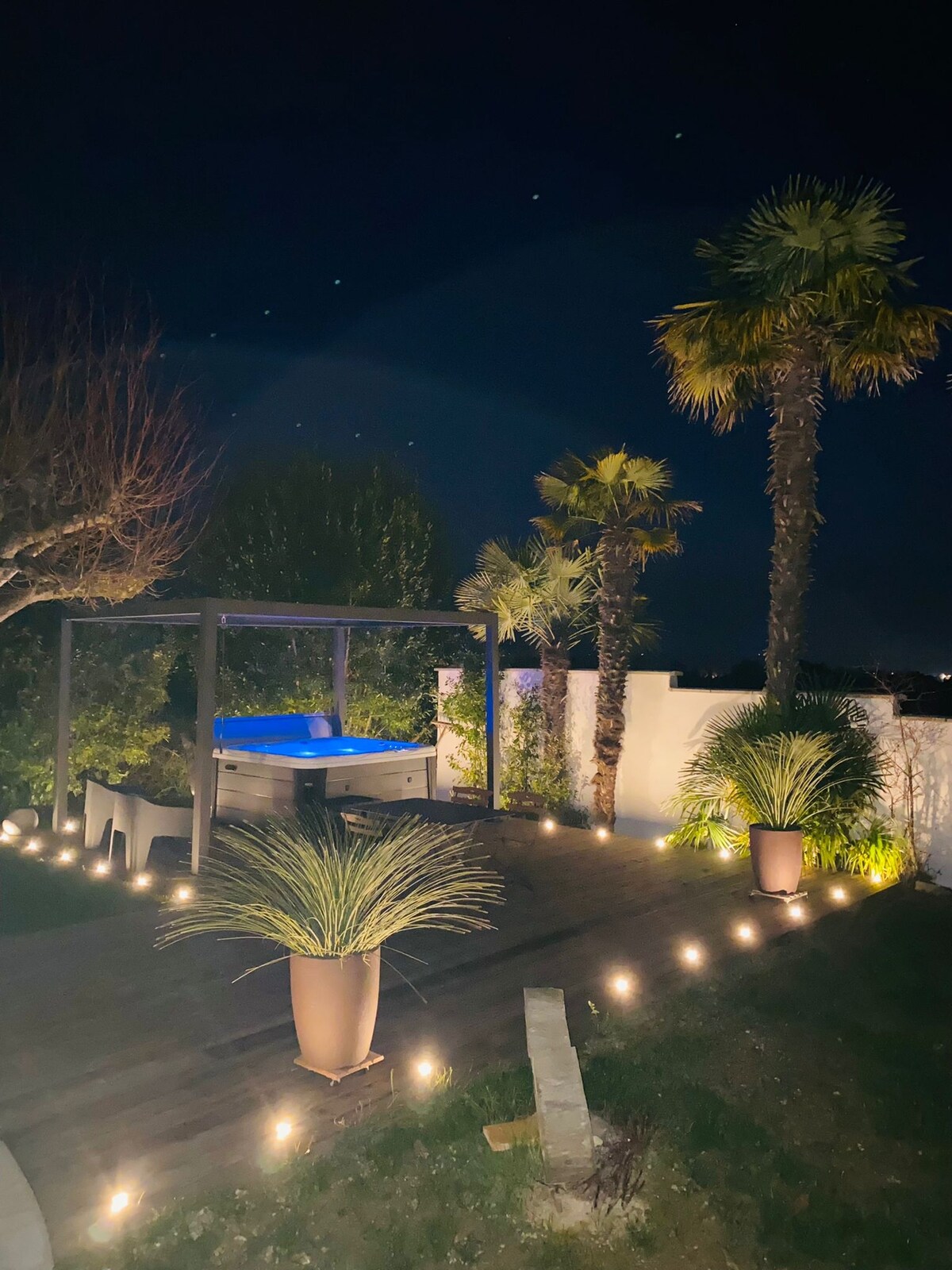 The outdoor area features a private jacuzzi, surrounded by lush palm trees and illuminated by soft outdoor lighting. Wooden decking creates a cozy atmosphere, while planters with greenery add texture to the serene setting at night.