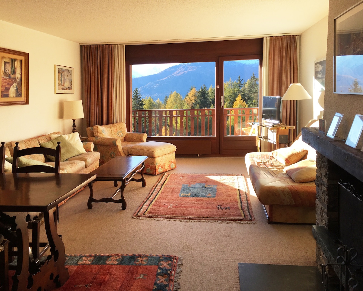 A spacious living area features large windows offering unobstructed views of the mountains. Two comfortable sofas and a cozy seating area are positioned around a central coffee table. A fireplace sits against one wall, accompanied by light-colored drapes and framed artwork.