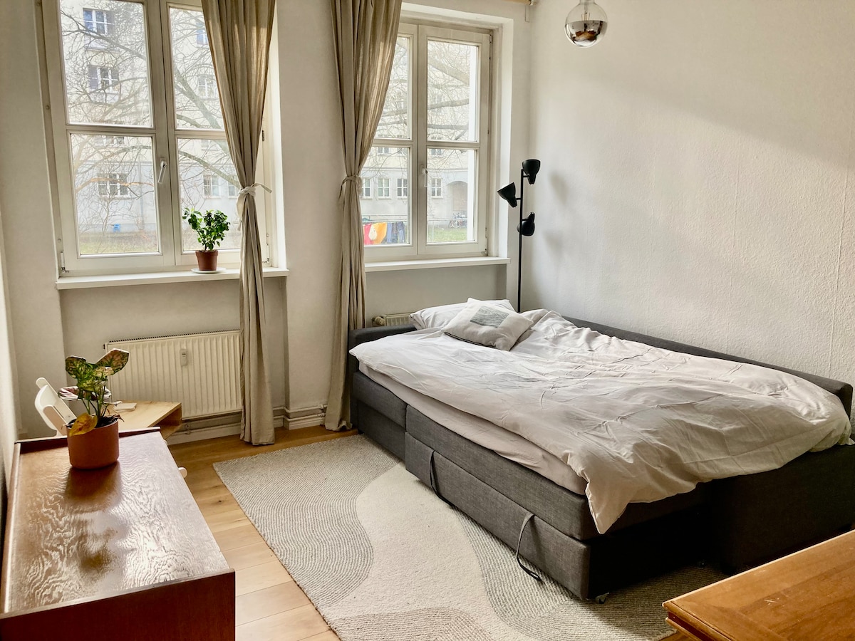 The living room features a comfortable sofa that converts into a double bed, topped with a real mattress. Natural light fills the space through large windows dressed in light curtains, with a small potted plant on a side table and a wooden dresser in view.