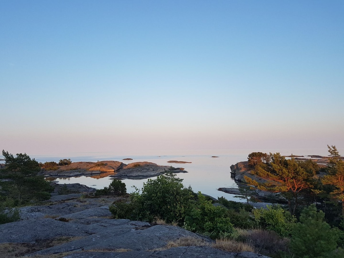 A serene coastal landscape features smooth, bare rock formations leading towards calm waters. The horizon is marked by scattered islands,. Light hues of blue and pink in the sky create a peaceful atmosphere, complemented by patches of greenery along the shore.